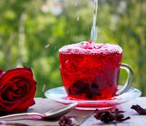 Clear cup of vibrant red hibiscus tea being poured, with a rose and hibiscus petals on the side.